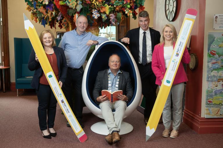 2 Men and 2 Women Standing holding pencils and one man sitting in egg chair reading