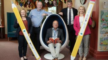 2 Men and 2 Women Standing holding pencils and one man sitting in egg chair reading